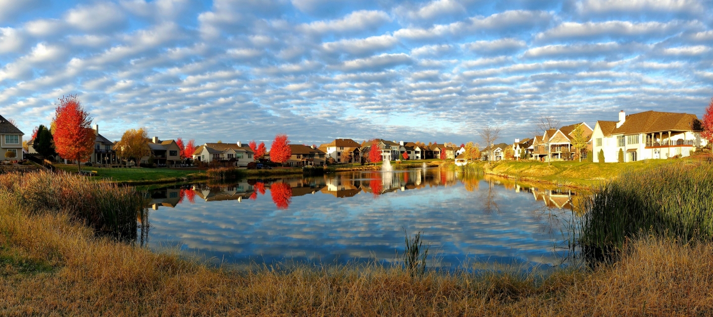 Panoramic image across a lake surrounded by houses and fall colored trees with a lightly clouded sky