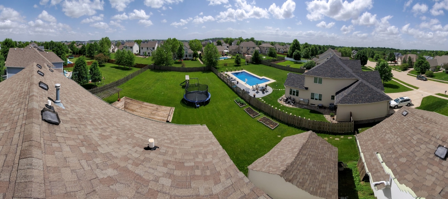 A panoramic image taken on top of a house roof overlooking multiple backyards -- one with a pool and another with a trampoline. The grass is vibrantly green and the sky is bright blue.